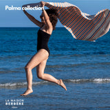 Frau springt am Meer und schwingt eine orange-blau gestreifte Palma-Fouta
